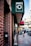 A city street scene with a brick building and a dark green awning featuring a laundry machine illustration. Below the awning, there is a glass door with a pull sign, and a man is walking on the sidewalk holding a coffee cup. The background includes urban elements like a street sign, bicycles, and parked vehicles.