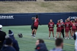 A group of female athletes in matching burgundy sports uniforms are on a field, with one of them joyfully jumping and celebrating with a coach or fellow team member. The scene captures a moment of camaraderie and excitement. In the background, other team members are standing in a line, some holding sports equipment.