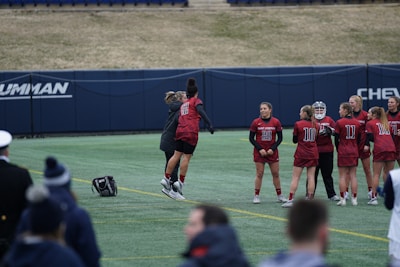 A group of female athletes in matching burgundy sports uniforms are on a field, with one of them joyfully jumping and celebrating with a coach or fellow team member. The scene captures a moment of camaraderie and excitement. In the background, other team members are standing in a line, some holding sports equipment.