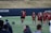 A group of female athletes in matching burgundy sports uniforms are on a field, with one of them joyfully jumping and celebrating with a coach or fellow team member. The scene captures a moment of camaraderie and excitement. In the background, other team members are standing in a line, some holding sports equipment.