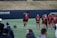 A group of female athletes in matching burgundy sports uniforms are on a field, with one of them joyfully jumping and celebrating with a coach or fellow team member. The scene captures a moment of camaraderie and excitement. In the background, other team members are standing in a line, some holding sports equipment.