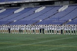 A group of athletes in matching white and dark-colored uniforms are standing in a line on a sports field. The background shows empty blue stadium seats and a wall with names inscribed on it.