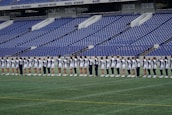 A group of athletes in matching white and dark-colored uniforms are standing in a line on a sports field. The background shows empty blue stadium seats and a wall with names inscribed on it.
