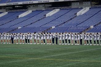 A group of athletes in matching white and dark-colored uniforms are standing in a line on a sports field. The background shows empty blue stadium seats and a wall with names inscribed on it.