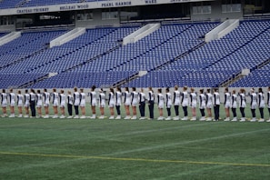 A group of athletes in matching white and dark-colored uniforms are standing in a line on a sports field. The background shows empty blue stadium seats and a wall with names inscribed on it.