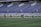 A group of athletes in matching white and dark-colored uniforms are standing in a line on a sports field. The background shows empty blue stadium seats and a wall with names inscribed on it.