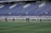 A group of athletes in matching white and dark-colored uniforms are standing in a line on a sports field. The background shows empty blue stadium seats and a wall with names inscribed on it.