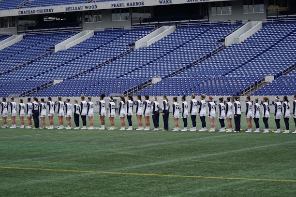 A group of athletes in matching white and dark-colored uniforms are standing in a line on a sports field. The background shows empty blue stadium seats and a wall with names inscribed on it.