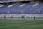 A group of athletes in matching white and dark-colored uniforms are standing in a line on a sports field. The background shows empty blue stadium seats and a wall with names inscribed on it.