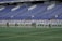 A group of athletes in matching white and dark-colored uniforms are standing in a line on a sports field. The background shows empty blue stadium seats and a wall with names inscribed on it.