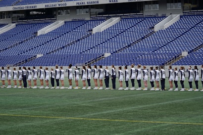 A group of athletes in matching white and dark-colored uniforms are standing in a line on a sports field. The background shows empty blue stadium seats and a wall with names inscribed on it.