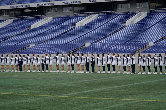 A group of athletes in matching white and dark-colored uniforms are standing in a line on a sports field. The background shows empty blue stadium seats and a wall with names inscribed on it.