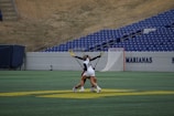 Two athletes are engaged in a lacrosse match on a green field with stadium seating in the background. The athlete on the left, wearing a white jersey with the number 54, is challenging another opponent for control of the ball in mid-action. Lacrosse sticks are extended and the players appear focused and competitive.