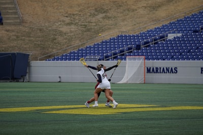 Two athletes are engaged in a lacrosse match on a green field with stadium seating in the background. The athlete on the left, wearing a white jersey with the number 54, is challenging another opponent for control of the ball in mid-action. Lacrosse sticks are extended and the players appear focused and competitive.