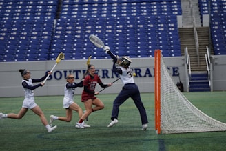 A lacrosse game taking place in an outdoor stadium with blue seats in the background. Four female players are in action; one in a red uniform is attempting to score, while the goalkeeper in a white uniform with dark sleeves is lunging to make a save. Two other players in white uniforms are nearby.