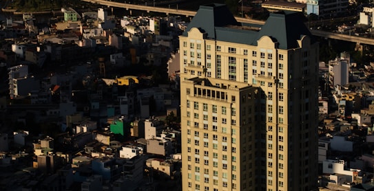 Tall residential or commercial building with a blue and beige facade dominates the foreground, surrounded by a densely packed urban landscape. The contrast between the well-lit building and the dimly lit surrounding area highlights the structure's prominence. Various smaller structures, possibly houses or low-rise buildings, are scattered around, creating a sense of urban density.