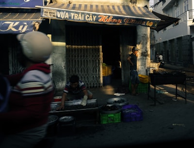 A street vendor is setting up goods for sale outside a fruit shop. Various containers and crates are scattered around, with one man wearing gloves handling merchandise, possibly preparing or arranging it. Another man stands nearby, observing or waiting. The shop has an awning with signage, and a person rides by on a motorcycle.