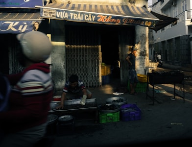 A street vendor is setting up goods for sale outside a fruit shop. Various containers and crates are scattered around, with one man wearing gloves handling merchandise, possibly preparing or arranging it. Another man stands nearby, observing or waiting. The shop has an awning with signage, and a person rides by on a motorcycle.