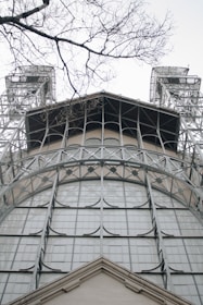 A detailed view of a metal-framed architectural structure, with a focus on intricate geometric patterns. The structure is flanked by two tall towers, and in the foreground, bare tree branches extend across the scene.
