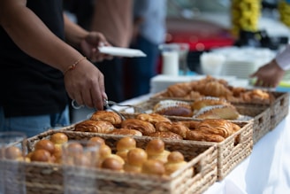 A selection of freshly baked pastries including croissants and other bread rolls arranged in woven baskets on a table. A person is serving themselves using tongs, while another person stands nearby. The setting suggests a buffet or self-service area with plates and glasses visible in the background.