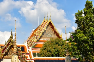 A traditional Thai temple roof adorned with intricate golden decorations against a backdrop of a clear blue sky. The roof's vivid colors of red and gold are complemented by lush green trees nearby. The architectural design features ornate spires and detailed patterns.