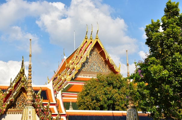 A traditional Thai temple roof adorned with intricate golden decorations against a backdrop of a clear blue sky. The roof's vivid colors of red and gold are complemented by lush green trees nearby. The architectural design features ornate spires and detailed patterns.