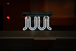 A brightly glowing neon sign with the word 'JUUL' is displayed against a dimly lit background. Above the sign, there is a warning label indicating the presence of nicotine and its addictive nature.