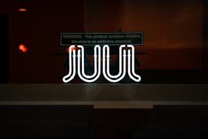 A brightly glowing neon sign with the word 'JUUL' is displayed against a dimly lit background. Above the sign, there is a warning label indicating the presence of nicotine and its addictive nature.
