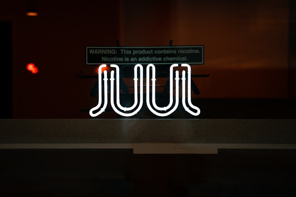 A brightly glowing neon sign with the word 'JUUL' is displayed against a dimly lit background. Above the sign, there is a warning label indicating the presence of nicotine and its addictive nature.