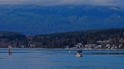 A serene lake view with a small boat on the water, surrounded by dense forested hills and mountains in the background. There are houses along the shoreline, and a navigation sign is visible in the water.