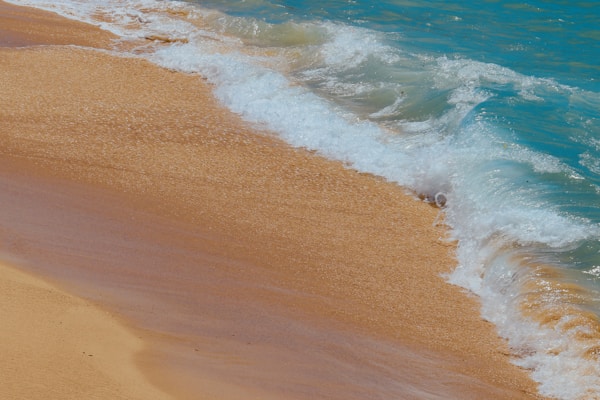 Gentle waves of a turquoise sea wash over a golden sandy beach, creating a delicate foam as the water retreats. The shoreline curves slightly, emphasizing the vibrant contrast between the blue ocean and the warm sand.