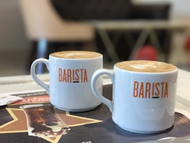 Two white mugs with 'BARISTA' written on them contain lattes with light-brown foam art. They are placed on a table with a magazine or pamphlet featuring a large star and a beverage image. The background is slightly blurred, suggesting a cozy, café-like setting.
