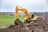 A large, yellow excavator is actively digging into a mound of earth in a rural landscape. The background features expansive fields and distant trees under a cloudy sky. A white vehicle is visible in the distance, parked near the fields.