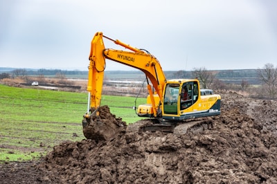 A large, yellow excavator is actively digging into a mound of earth in a rural landscape. The background features expansive fields and distant trees under a cloudy sky. A white vehicle is visible in the distance, parked near the fields.
