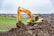 A large, yellow excavator is actively digging into a mound of earth in a rural landscape. The background features expansive fields and distant trees under a cloudy sky. A white vehicle is visible in the distance, parked near the fields.