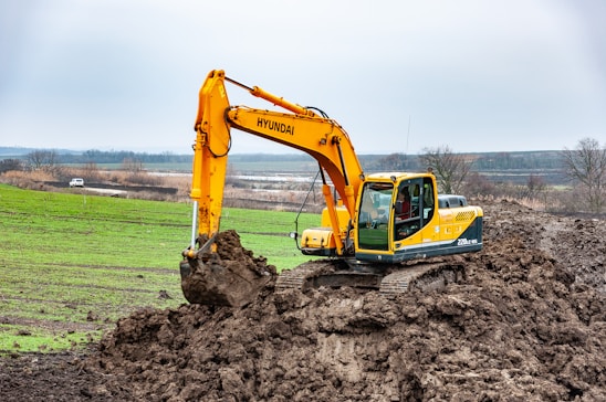 A large, yellow excavator is actively digging into a mound of earth in a rural landscape. The background features expansive fields and distant trees under a cloudy sky. A white vehicle is visible in the distance, parked near the fields.