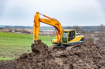 A large, yellow excavator is actively digging into a mound of earth in a rural landscape. The background features expansive fields and distant trees under a cloudy sky. A white vehicle is visible in the distance, parked near the fields.