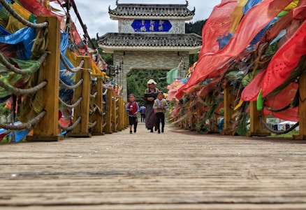 A wooden bridge adorned with colorful, flowing fabrics on either side. Three people, two children with an adult, walk across the bridge towards a traditional architectural structure in the background. The setting is outdoors with a natural landscape visible beyond the wooden posts and ropes lining the bridge.