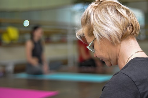 A blonde woman with glasses is seen in profile, focusing on something out of the frame. The background is blurred, with another person seated on a yoga mat indoors. The overall setting suggests a calm environment, likely a yoga or relaxation class.