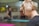 A blonde woman with glasses is seen in profile, focusing on something out of the frame. The background is blurred, with another person seated on a yoga mat indoors. The overall setting suggests a calm environment, likely a yoga or relaxation class.