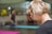 A blonde woman with glasses is seen in profile, focusing on something out of the frame. The background is blurred, with another person seated on a yoga mat indoors. The overall setting suggests a calm environment, likely a yoga or relaxation class.