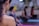A close-up of a person's shoulder and head, with their hair tied back, in the foreground. In the background, several other people are seated on yoga mats in a blurred focus, suggesting a yoga or meditation class setting.