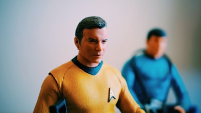 A model of a character in a yellow-orange uniform with a black collar and an insignia badge on the chest. The model has short dark hair and is positioned in focus. In the background, another character appears out of focus, wearing a blue uniform and also with dark hair.