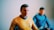 A model of a character in a yellow-orange uniform with a black collar and an insignia badge on the chest. The model has short dark hair and is positioned in focus. In the background, another character appears out of focus, wearing a blue uniform and also with dark hair.