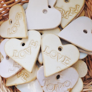 A collection of heart-shaped ceramic tags with words like 'LOVE' and different textures, arranged in a wicker basket. The hearts have small holes near the top, possibly for hanging.