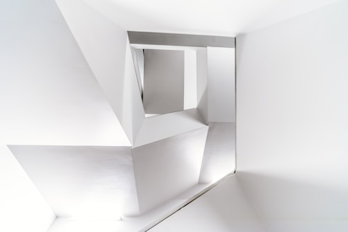 A modern architectural interior with clean, geometric lines and sharp angles in a monochromatic color scheme. The structure features an arrangement of triangular and rectangular shapes creating an abstract and minimalist design.