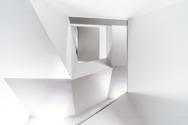 A modern architectural interior with clean, geometric lines and sharp angles in a monochromatic color scheme. The structure features an arrangement of triangular and rectangular shapes creating an abstract and minimalist design.