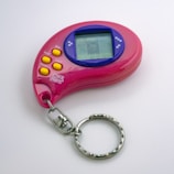 A pink handheld electronic toy with a digital screen and several yellow buttons, including options for mode, accept, cancel, and reverse. The device is shaped in a curved design with a keychain attached.