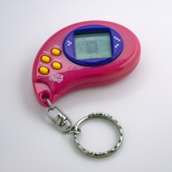 A pink handheld electronic toy with a digital screen and several yellow buttons, including options for mode, accept, cancel, and reverse. The device is shaped in a curved design with a keychain attached.