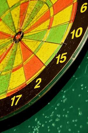 A close-up view of a colorful dartboard showing sections numbered from 6 to 17. The dartboard features bright yellow, red, and green sections with distinct black outlines. It is set against a green background with markings, suggesting previous gameplay.